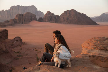 Young couple sitting on desert rock with cat at sunset - PCLF00572