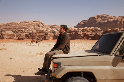 Nachdenklicher junger Mann sitzt auf einem Auto in der Wüste - PCLF00588
