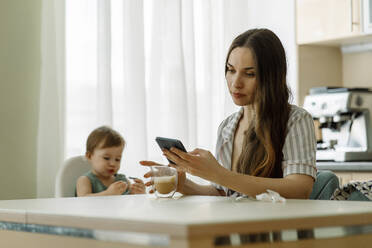 Mutter verwendet Smartphone mit Tochter sitzt auf dem Tisch zu Hause - IEF00412