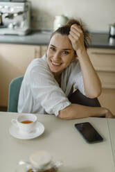Lächelnde Frau sitzt mit Smartphone und Tee am Tisch - IEF00418