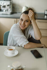 Smiling woman sitting on table with smart phone and tea - IEF00418