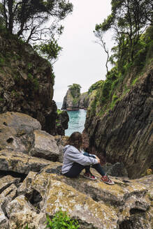 Junge Frau schaut auf das Meer und sitzt auf einer Felsformation - RSGF00903