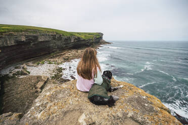 Junge Frau mit Blick auf das Meer auf einem Felsen sitzend mit Hund - RSGF00905