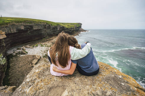 Frau mit Arm um Freund sitzt auf einem Felsen und schaut aufs Meer - RSGF00908