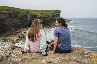 Young friends talking to each other sitting on rock near beach - RSGF00909