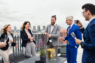 Eine große Gruppe fröhlicher Geschäftsleute feiert auf einer Dachterrasse in der Stadt eine Party und öffnet eine Flasche Champagner. - HPIF18527