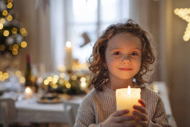 Vorderansicht eines fröhlichen kleinen Mädchens, das zu Weihnachten in einem Haus steht und eine Kerze hält. - HPIF18685