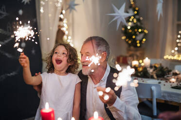 Portrait of senior grandfather with small granddaughter indoors at Christmas, sitting at table with sparklers. - HPIF18736