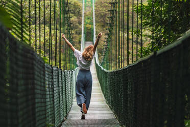 Carefree woman with arms raised spending leisure time walking on suspension bridge - RSGF00924