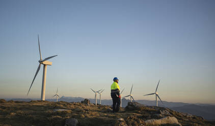 Ingenieur, der den Sonnenuntergang inmitten von Windkraftanlagen betrachtet - SNF01683