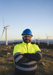 Smiling engineer with arms crossed standing in front of wind turbines - SNF01687