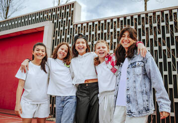 Lächelnde Teenager-Mädchen, die vor einer Mauer auf einem Spielplatz stehen und sich umarmen - MDOF01161