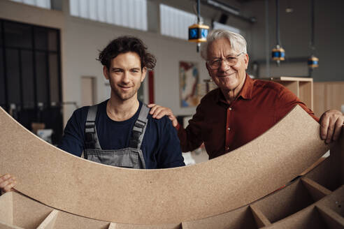 Lächelnder Tischler mit Geschäftsmann, der ein Holzbrett in der Werkstatt hält - JOSEF19416