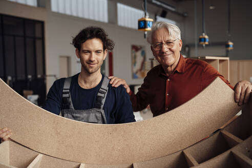 Smiling carpenter with businessman holding wooden plank at workshop - JOSEF19416