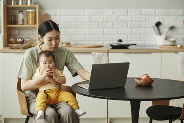 Freelancer mother using laptop with daughter sitting on lap at home - KPEF00004