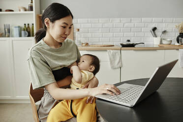 Smiling freelancer breastfeeding daughter and using laptop at home - KPEF00007