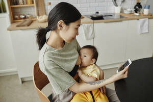 Mutter, die ihre Tochter stillt und zu Hause ein Smartphone benutzt - KPEF00008