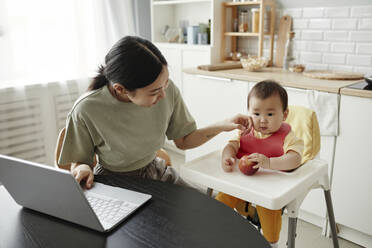 Freelancer mother wiping daughter's cheek sitting on seat at home - KPEF00011