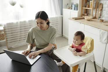 Freelancer mother working on laptop by daughter at home - KPEF00013
