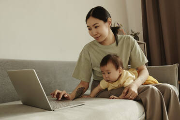 Freelancer mother using laptop sitting on sofa with daughter at home - KPEF00018