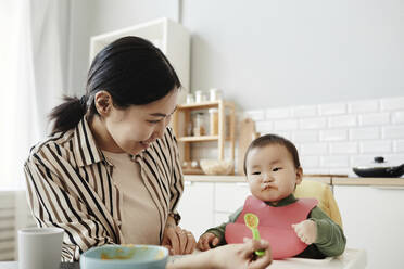 Lächelnde Frau füttert ihre Tochter zu Hause mit Fruchtpüree - KPEF00062