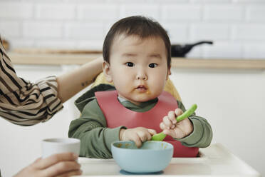 Cute daughter with bowl and spoon sitting in high chair at home - KPEF00063