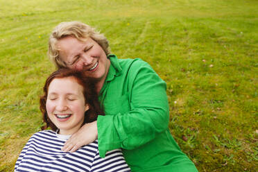 Happy mother embracing daughter sitting on grass - IHF01357