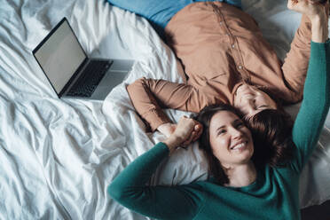 Happy lesbian couple lying down on bed with laptop at home - JOSEF19456
