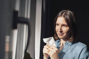 Thoughtful woman holding paper napkin near window at home - JOSEF19465