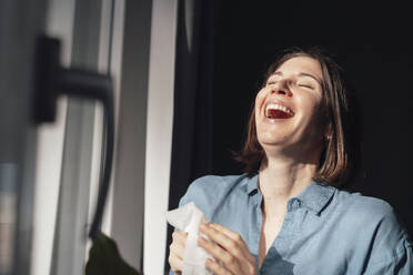 Cheerful woman laughing near window at home - JOSEF19466