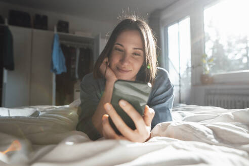 Smiling woman using smart phone lying down on bed at home - JOSEF19474