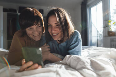 Curious lesbian couple sharing smart phone lying on bed at home - JOSEF19475