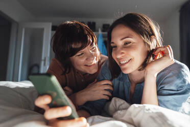Lächelndes lesbisches Paar mit Smartphone auf dem Bett zu Hause - JOSEF19476