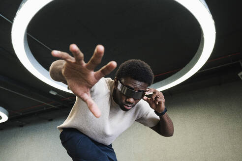 Young man with futuristic looking through cyber glasses under modern ring lamp reaching out hand - PNAF05293