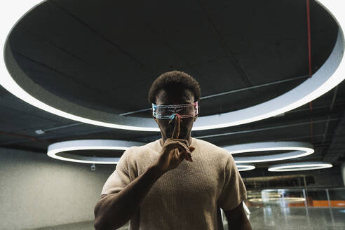 Young man with futuristic cyber glasses under modern ring lamp putting forefinger on lips - PNAF05295