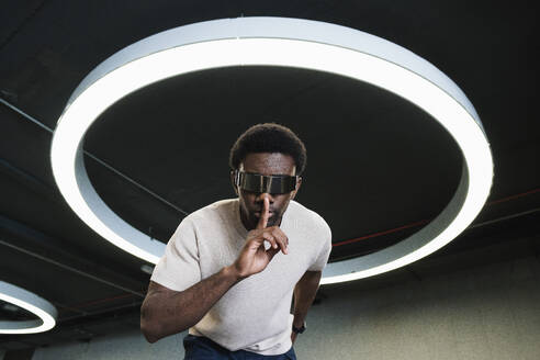 Young man with futuristic cyber glasses under modern ring lamp putting forefinger on lips - PNAF05297