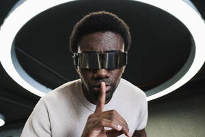 Young man with futuristic cyber glasses under modern ring lamp putting forefinger on lips - PNAF05299