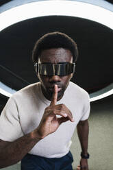 Young man with futuristic cyber glasses under modern ring lamp putting forefinger on lips - PNAF05300