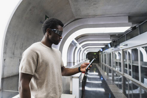 Man wearing futuristic glasses standing at metro station using smartphone - PNAF05312