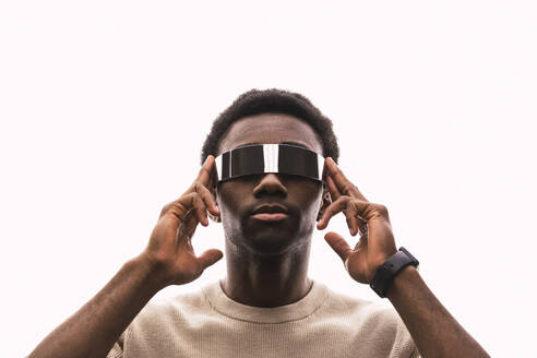 Cool young man wearing cyber glasses against white background putting fingers on temples - PNAF05338