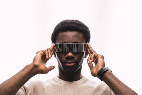 Cool young man wearing cyber glasses against white background putting fingers on temples - PNAF05343