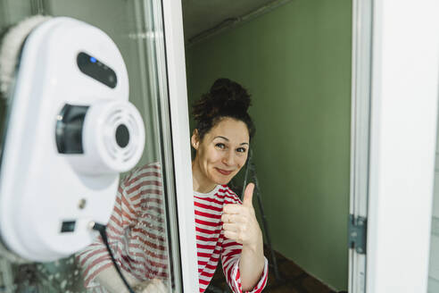 Glückliche Frau zeigt Daumen hoch Geste und Reinigung Fenster mit Roboter Waschmaschine - IEF00447