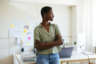 Happy thoughtful freelancer standing with arms crossed in home office - EBSF03418