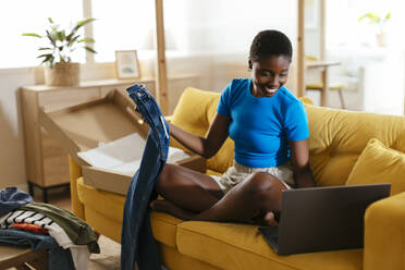 Happy woman checking clothes and using laptop at home - EBSF03432