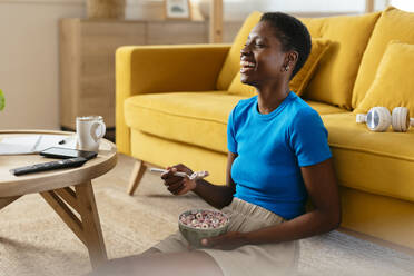 Fröhliche junge Frau isst Müsli und sieht zu Hause fern - EBSF03437