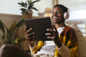 Fröhliche junge Frau mit drahtlosen Kopfhörern, die zu Hause einen Film auf einem Tablet-PC anschaut - EBSF03450