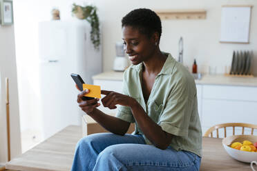 Smiling young woman making payment through credit card on mobile phone at home - EBSF03461