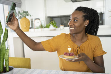 Eine fröhliche Frau macht ein Selfie mit ihrem Smartphone, während sie eine köstliche Himbeertorte in ihrer Wohnung hält - SVKF01393