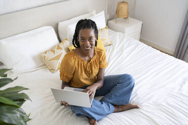 Smiling freelancer sitting with laptop on bed - SVKF01397
