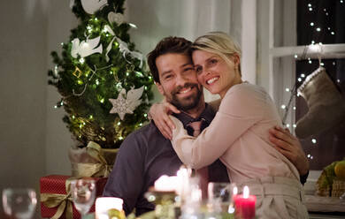 Vorderansicht eines glücklichen Paares, das drinnen am Tisch sitzt und Weihnachten feiert und sich umarmt. - HPIF18758
